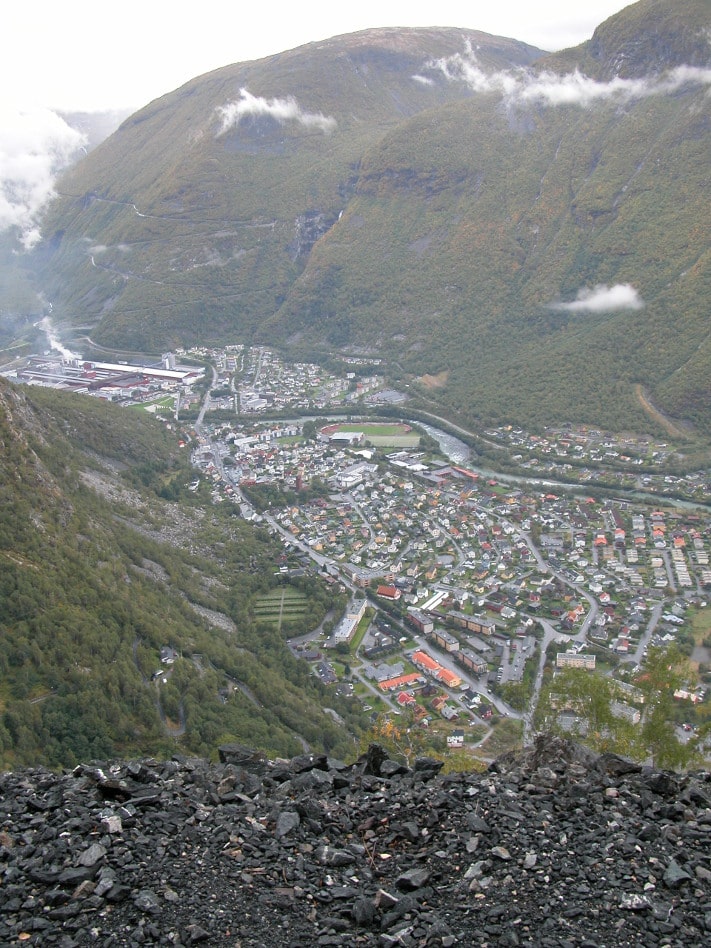 Utsikt over dagens Farnes (Øvre Årdal) sett fra gruvetippen i Blåberget. Der nede lå smeltehytten og gruvebyen. Til venstre i disen ser vi litt av den kjente veien opp til den 1469 meter høye Heirsnosi. Den smale veien hadde 42 krappe hårnålssvinger opp til 1000 meter. Veien ble bygget av Hydro og var ferdig i 1912 som anleggsvei til starten av utbyggingen av Tyin-fallene. Etter framføringen til Tyin i 1934 var den lenge Årdals eneste veiforbindelse med utenverdenen. I 1965 ble den helårsvei etter omlegging og utbygging. Den gamle veien står på Veivesenets verneplan. Fra 1000-meteren og ned til kraftstasjonen i fjellet nede i bygda, går kraftverkets trykksjakt i tunnel. Foto: Helge Haugen